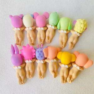 May include: A collection of whimsical figurines, each featuring a nude body with a variety of colorful, cartoonish headpieces. The heads include bunny ears, mouse ears, and other abstract shapes in shades of pink, yellow, green, and purple. The figurines are arranged closely together.