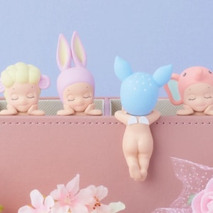 May include: A collection of four small figurines, each with a different animal-themed head covering, resting on a pink box. The figurines include a sheep, a bunny, a deer, and an elephant. The background is a soft blue.
