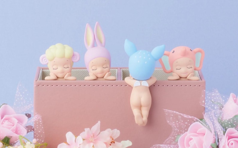 May include: A collection of four small figurines, each with a different animal-themed head covering, resting on a pink box. The figurines include a sheep, a bunny, a deer, and an elephant. The background is a soft blue.