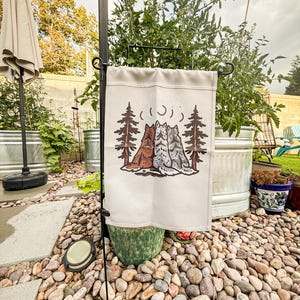 May include: A rectangular off-white garden flag hanging on a black metal stand. The flag features a graphic illustration of three wolves sitting back-to-back between two tall pine trees, with the word "BLOOMING" and moon phases printed above.