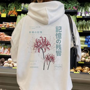 May include: A white hooded sweatshirt featuring a graphic print on the back. The design includes two red spider lily flowers, Japanese text, and English phrases like Shadow Bloom Memories, Archive 07, and Fade Signal in a minimalist layout.