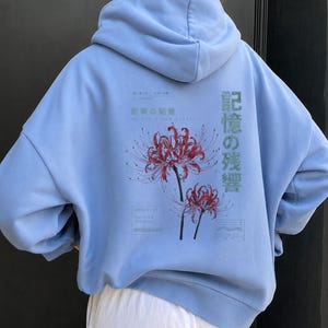 May include: A light blue hooded sweatshirt featuring a graphic on the back with two red spider lily flowers. The design includes Japanese text, specifically 記憶の残響 and 影の記憶, along with small rectangular boxes containing additional text.