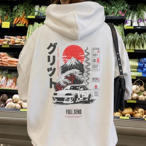 May include: A white hooded sweatshirt featuring a graphic on the back with a red sun, a mountain, a wave, and a white car. The text includes Japanese characters, 'GRIP CONTROL DESTINY', 'FULL SEND', and various technical symbols and coordinates.