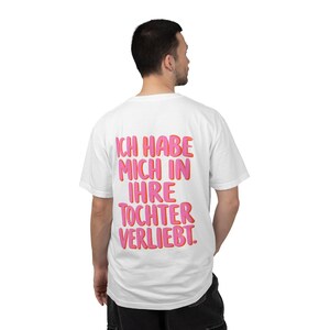 Puede incluir: Una camiseta blanca de manga corta con texto rosa intenso en la espalda que dice "ICH HABE MICH IN IHRE TOCHTER VERLIEBT." Las letras tienen un contorno naranja sutil y la prenda se muestra desde una perspectiva trasera sobre fondo liso.