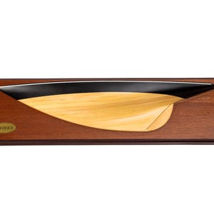 Könnte beinhalten: Ein Holzbootsmodell mit schwarzer Oberseite und hellem Holzrumpf, montiert auf einer rechteckigen, dunklen Holzbasis. Das Modell hat ein schlankes, geschwungenes Design, und ein kleines, ovales Emblem ist auf der Basis sichtbar.