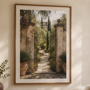 Può includere: Una stampa artistica incorniciata che raffigura un cancello in ferro battuto aperto che conduce a un sentiero in pietra in un giardino mediterraneo, con pilastri in pietra consumata, vasi in terracotta e cipressi sotto un cielo luminoso.