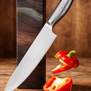 May include: A stainless steel chef's knife with a silver blade and a patterned handle. A red bell pepper is cut in half, with a green stem, and is next to the knife. The knife is next to a wooden box.