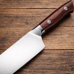 May include: Close-up of a stainless steel chef's knife with a dark brown wooden handle. The handle is secured with two silver rivets. The blade is sharp and angled, reflecting the light. The knife rests on a wooden surface.
