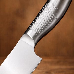 May include: Close-up of a stainless steel kitchen knife. The blade is shiny and sharp, with a curved handle featuring a textured pattern of small, evenly spaced dimples. The background is a blurred wooden surface.