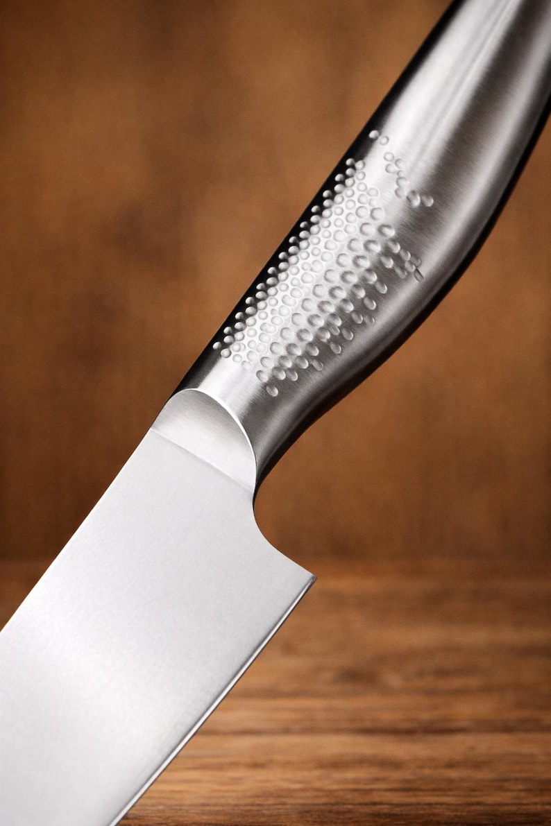 May include: Close-up of a stainless steel kitchen knife. The blade is shiny and sharp, with a curved handle featuring a textured pattern of small, evenly spaced dimples. The background is a blurred wooden surface.