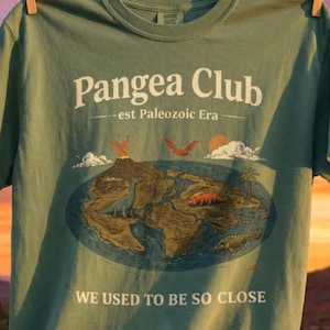 Peut inclure: T-shirt vert sauge avec le texte "Pangea Club - est Paleozoic Era" et un graphique du supercontinent Pang&eacute;e avec des dinosaures. Le t-shirt porte &eacute;galement le texte "WE USED TO BE SO CLOSE". Le t-shirt est suspendu &agrave; une corde &agrave; linge avec un arri&egrave;re-plan d&eacute;sertique.
