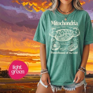 May include: Light green t-shirt with the word "Mitochondria" and a diagram of a cell's mitochondria. The shirt also has the text "Powerhouse of The Cell!" and other scientific terms. The shirt is worn with denim shorts.