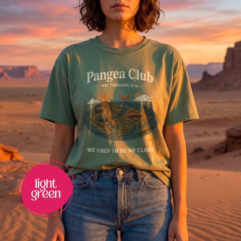 Peut inclure: T-shirt vert clair avec le texte "Pangea Club - est Paleozoic Era" et la phrase "WE USED TO BE SO CLOSE". Le t-shirt pr&eacute;sente un graphisme d'un paysage pr&eacute;historique. Le mod&egrave;le porte un jean bleu.
