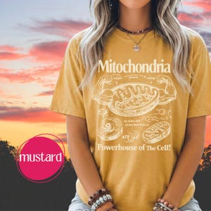 May include: Mustard-colored t-shirt with a white graphic of a cell's mitochondria and the text "Mitochondria Powerhouse of The Cell!". The shirt features a diagram of the cell's components and the words "oxygen", "glucose", and "energy".