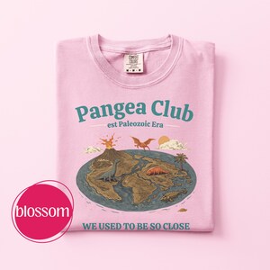 Peut inclure: T-shirt rose clair avec un motif graphique du Pangea Club. Le design pr&eacute;sente une carte du supercontinent avec des dinosaures, un volcan et le texte "WE USED TO BE SO CLOSE". Le t-shirt a un col rond et une &eacute;tiquette Comfort Colors.