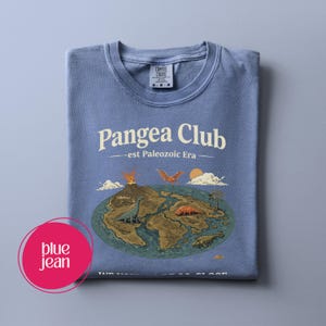 Peut inclure: T-shirt bleu jean pli&eacute; avec l'inscription "Pangea Club - est Paleozoic Era". Le motif repr&eacute;sente la Terre avec des dinosaures, des volcans et des palmiers. Le t-shirt porte une &eacute;tiquette Comfort Colors.