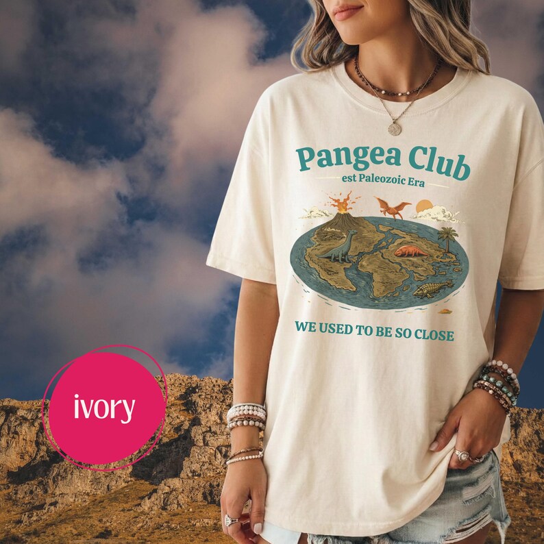 Peut inclure: T-shirt ivoire avec un motif du Pangea Club, repr&eacute;sentant une carte du supercontinent avec des dinosaures, un volcan et le texte "WE USED TO BE SO CLOSE." Le t-shirt est de coupe ample.