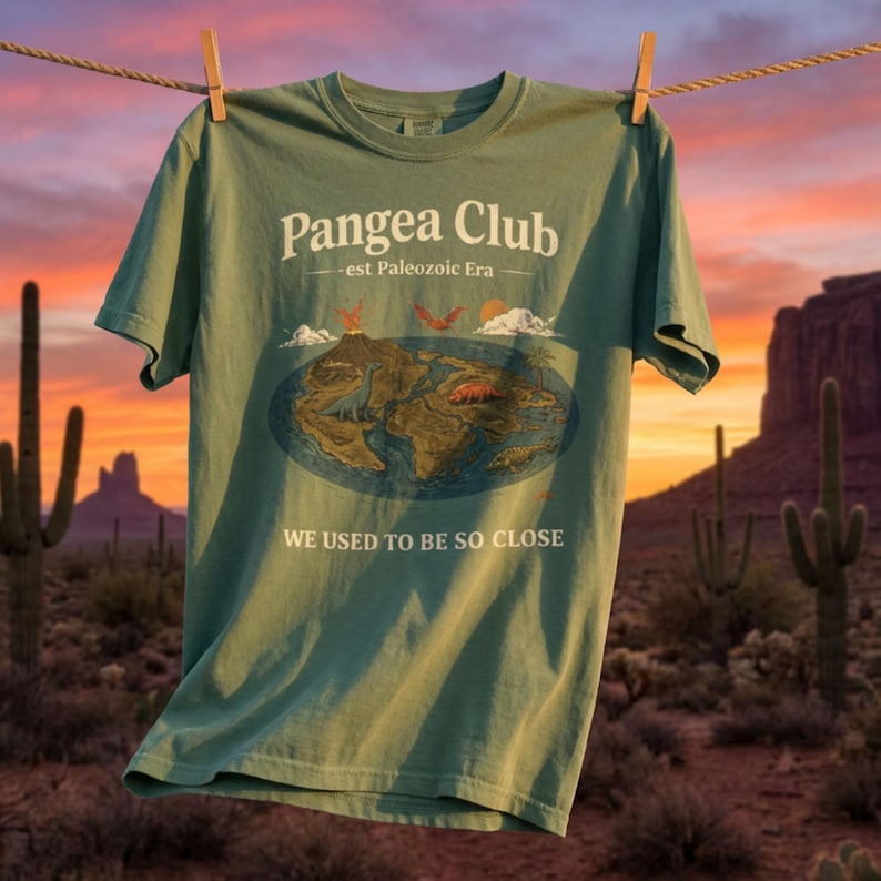Peut inclure: T-shirt vert sauge avec le texte "Pangea Club - est Paleozoic Era" et un graphique du supercontinent Pang&eacute;e avec des dinosaures. Le t-shirt porte &eacute;galement le texte "WE USED TO BE SO CLOSE". Le t-shirt est suspendu &agrave; une corde &agrave; linge avec un arri&egrave;re-plan d&eacute;sertique.