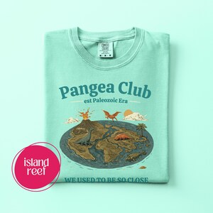 Peut inclure: T-shirt vert clair avec l'inscription "Pangea Club" et "est Paleozoic Era". Le t-shirt pr&eacute;sente un dessin de la Terre avec des dinosaures, un volcan et le texte "WE USED TO BE SO CLOSE". Le t-shirt a un col rond.