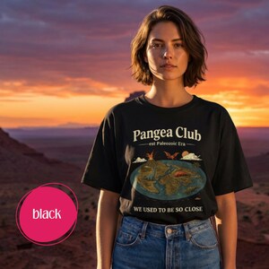 Peut inclure: T-shirt noir avec l'inscription "Pangea Club - est Paleozoic Era" et un dessin du supercontinent de la Pang&eacute;e. Le t-shirt porte &eacute;galement l'inscription "WE USED TO BE SO CLOSE". Le mod&egrave;le porte un jean bleu.