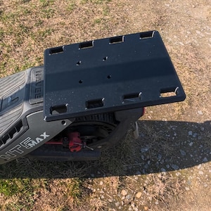 Könnte beinhalten: Schwarze Gepäckplattform an einem schwarz-grauen Elektroroller befestigt. Die Plattform hat mehrere rechteckige Aussparungen und Befestigungslöcher. Der Roller hat den Text "NTE MAX" an der Seite.