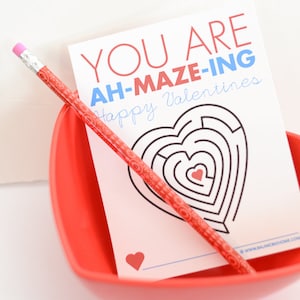 May include: A white card with the text "You are Ah-Mazing Happy Valentines" and a heart-shaped maze. A red pencil is lying on top of the card.