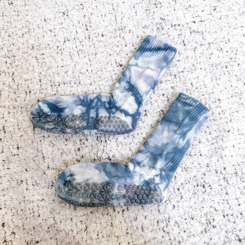 May include: A pair of tie-dye socks in shades of blue and white. The socks have a ribbed cuff and a non-slip grip sole. The tie-dye pattern is a mix of swirling and mottled designs. The socks are designed for comfort and style.