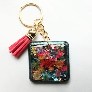 Op de afbeelding: Een vierkante sleutelhanger van hars met een donkergroenblauwe rand en een verzameling kleine, kleurrijke gedroogde bloemen erin verwerkt. De sleutelhanger heeft een goudkleurige metalen ring en een kort, felroze kwastje van imitatie-suède.