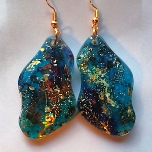 Op de afbeelding: Een paar hangende oorbellen met onregelmatige, golvende hars hangers in een doorschijnende blauwgroene kleur. De hangers zijn ingelegd met glinsterende gouden en iriserende vlokken die een textuurrijk uiterlijk creëren, voorzien van gouden haken.