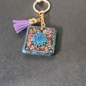 Op de afbeelding: Een vierkante sleutelhanger met een goudkleurige metalen ring en ketting. De hanger heeft een textuur met veelkleurige glitters en een centrale blauwe bloemvormige versiering. Een klein lichtpaars kwastje is aan de ketting bevestigd.