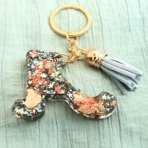 May include: A letter A keychain made of clear resin filled with multicolored star-shaped glitter and copper-colored metallic flakes. The letter is attached to a gold-toned metal split ring and a short, light gray suede leather tassel.
