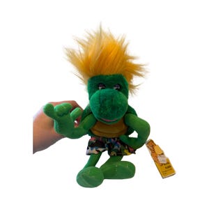 Peut inclure: Une peluche dinosaure verte avec une crête jaune et un short coloré. Le dinosaure a une expression amicale et lève une main. Une étiquette jaune est attachée au jouet.