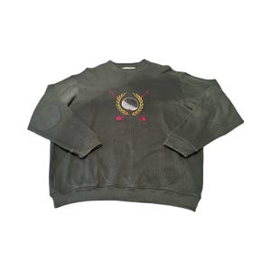 May include: A dark charcoal gray crewneck sweatshirt featuring long sleeves with circular elbow patches. The chest displays an embroidered graphic of a white golf ball centered within a yellow laurel wreath, flanked by two crossed red golf clubs.