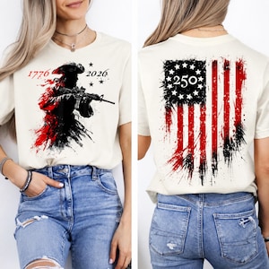 以下が含まれることがあります： 兵士がライフルを持っているグラフィックが特徴のクリーム色のTシャツ。前面には「1776」と「2026」の数字がプリントされています。背中には、数字「250」が付いた、使い古されたアメリカ国旗のデザインが施されています。