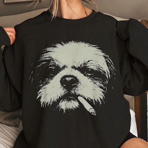 May include: A black crewneck sweatshirt featuring a large, light-colored graphic print of a shaggy dog face. The dog is depicted with a cigarette held in its mouth. The design is centered on the chest, showcasing a high-contrast, detailed illustration of the canine's fur texture and facial features against the dark fabric background.