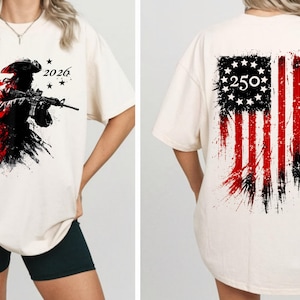 以下が含まれることがあります： クリーム色のTシャツ。ライフルを持った兵士のグラフィック、赤と黒のペイントスプラッター、数字の「1776」と「2026」が特徴です。背中には、数字「250」が付いた、使い古されたアメリカ国旗のデザインが表示されています。