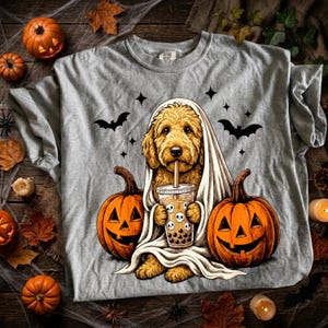 May include: A heather gray t-shirt featuring a graphic of a golden-colored dog draped in a white sheet like a ghost. The dog holds a bubble tea cup with skull icons. Two orange jack-o'-lanterns and black bats surround the central figure.