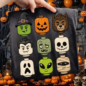 May include: A black t-shirt featuring a graphic print of nine stylised toy-style character heads arranged in a grid. The characters include a witch, a jack-o'-lantern, a werewolf, a vampire, a zombie, a skeleton, a ghost, an alien, and a mummy.