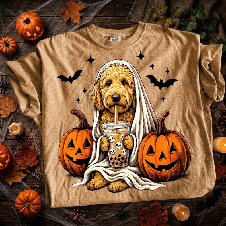 May include: A tan t-shirt featuring a golden doodle dog draped in a white sheet like a ghost, holding a plastic cup of boba tea with skull icons. The dog is flanked by two orange jack-o'-lanterns, with black bats and stars in the background.