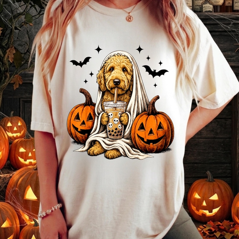 May include: A cream-colored t-shirt featuring a graphic of a golden-colored dog draped in a white sheet like a ghost, holding a bubble tea cup with skull designs, flanked by two orange jack-o'-lanterns and surrounded by black bat silhouettes.