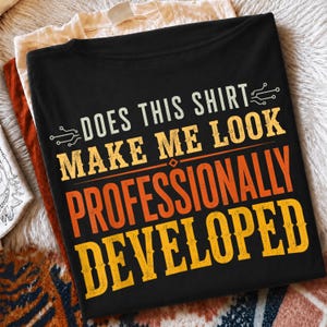 Peut inclure: Un t-shirt noir portant l'inscription "DOES THIS SHIRT MAKE ME LOOK PROFESSIONALLY DEVELOPED" en lettres jaunes et orange, agrémenté de petites icônes graphiques de circuits imprimés de chaque côté de la première ligne de texte.