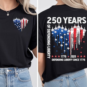 Könnte beinhalten: Schwarzes T-Shirt mit patriotischem Design. Die Vorderseite zeigt ein Herz aus weißen Flugzeugen, blauen Sternen und roten Streifen. Auf der Rückseite steht "250 YEARS OF DEFENDING LIBERTY" und "DEFENDING LIBERTY SINCE 1776".
