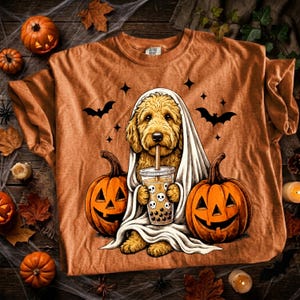 May include: An orange t-shirt featuring a graphic of a golden-colored dog draped in a white sheet like a ghost, holding a bubble tea cup with skull designs. The dog is positioned between two orange jack-o'-lanterns with black bats above.