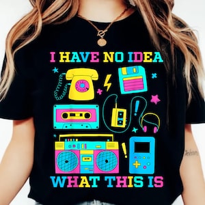 以下が含まれることがあります： 黒いTシャツ。レトロな電子機器のカラフルなグラフィックが特徴。電話、カセットテープ、フロッピーディスク、ラジカセなど。テキスト「I HAVE NO IDEA WHAT THIS IS」がグラフィックの上と下に明るい色で印刷されています。