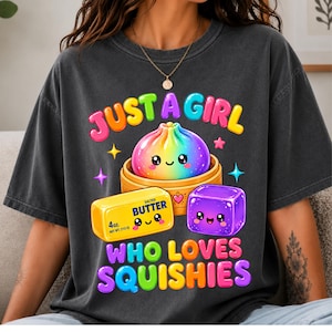 May include: A charcoal gray t-shirt featuring a colorful graphic with the text "JUST A GIRL WHO LOVES SQUISHIES". The design includes a rainbow-colored dumpling in a bamboo steamer, a yellow stick of 4oz salted butter, and a purple cube.