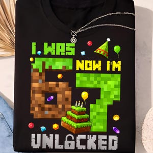 May include: Black t-shirt with a colorful graphic design. The design features the text "I WAS 6 NOW I'M 7 UNLOCKED" in pixelated font, along with a pixelated cake, balloons, and confetti. A silver necklace is visible.