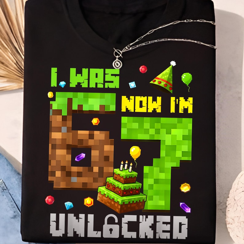 May include: Black t-shirt with a colorful graphic design. The design features the text "I WAS 6 NOW I'M 7 UNLOCKED" in pixelated font, along with a pixelated cake, balloons, and confetti. A silver necklace is visible.