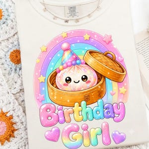 May include: A cream-colored t-shirt featuring a graphic of a smiling dumpling in a bamboo steamer wearing a party hat. A rainbow arches behind the dumpling, surrounded by stars and hearts. The text "Birthday Girl" appears in large, colorful letters.