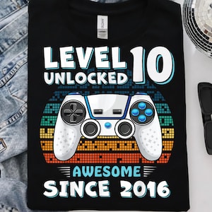 May include: A black t-shirt featuring a graphic print with the text "LEVEL 10 UNLOCKED" and "AWESOME SINCE 2016". The design includes a white video game controller centred over a retro-style sunset background composed of orange, yellow, and blue pixels.