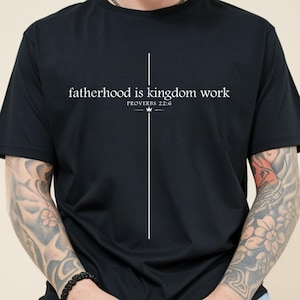 May include: Black t-shirt featuring the white text "fatherhood is kingdom work" and the Bible reference Proverbs 22:6. A white vertical line and crown design are centered on the shirt.